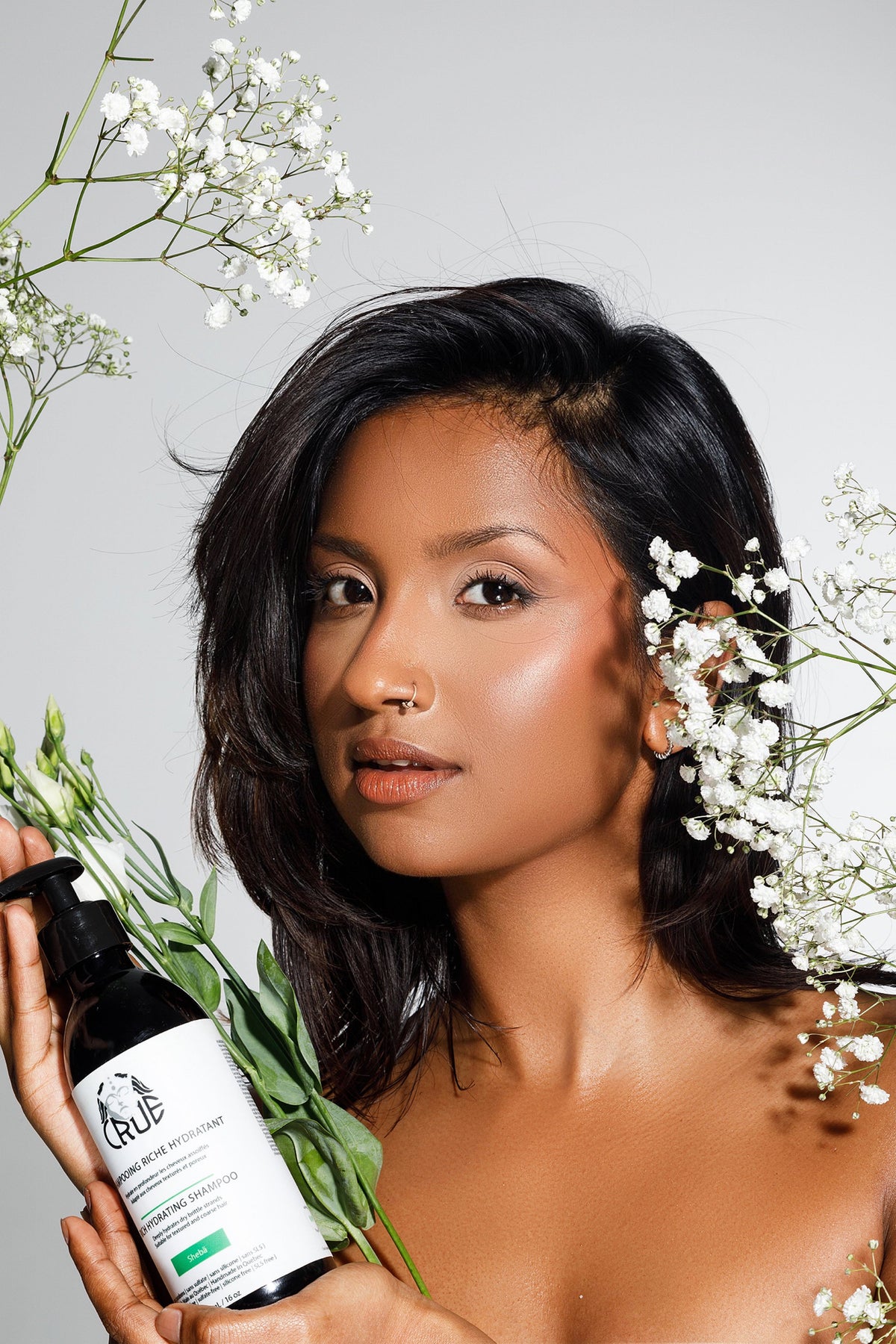 Femme aux cheveux brillants entourée de fleurs, flacon Crüe à la main